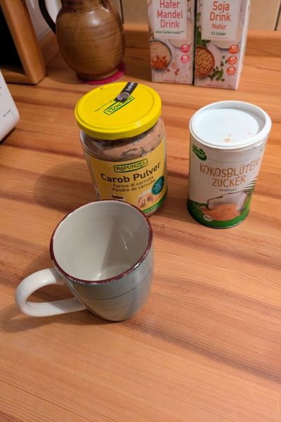 Auf einer hölzernen Arbeitsfläche steht eine Keramiktasse, dahinter ein Glas mit Carobpulver und ein Becher mit Kokosblütenzucker.