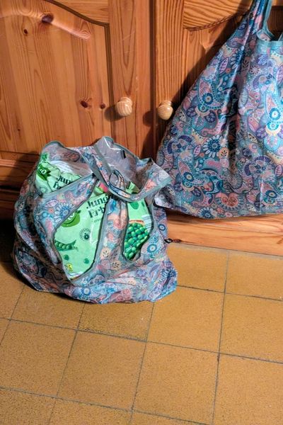 Auf braunen Fliesen steht ein Einkaufsbeutel in blau-rosa Mustern. Ein zweiter Einkaufsbeutel hängt an einem hölzernen Bauernschrank direkt dahinter.