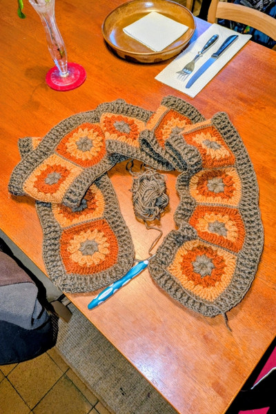 Auf einem Küchentisch liegt ein gehäkelter Schal. Das Muster sind aneinander gehäkelte Daisy-Grannys in herbstlichen Farben: braun, kürbis-gelb und rostrot.