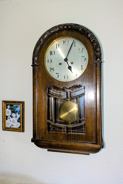 Ein alter, dunkel braun gebeizter Regulator ( Pendeluhr) hängt an der Wand, daneben hängt ein Bild von wilden Hundsrosen.