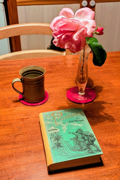 Auf einem tisch stehen eine Tasse Tee und eine Rose. Davor liegt ein Buch: "Die Pilgerreise" von John Bunyan.