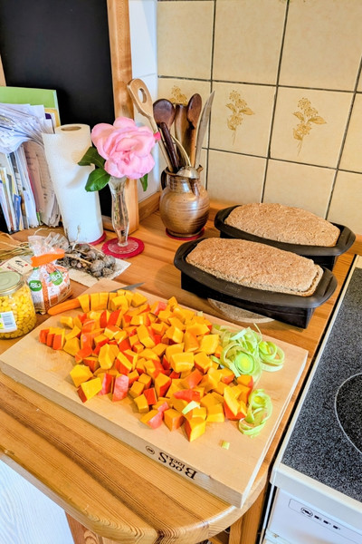Ein Schneidbrett mit Porree, Kürbiswürfel, Mais, Linsen- Zutaten für eine Kürbissuppe. Dahinter liegen zwei Laibe Brot und kühlen nach dem Backen weiter ab.