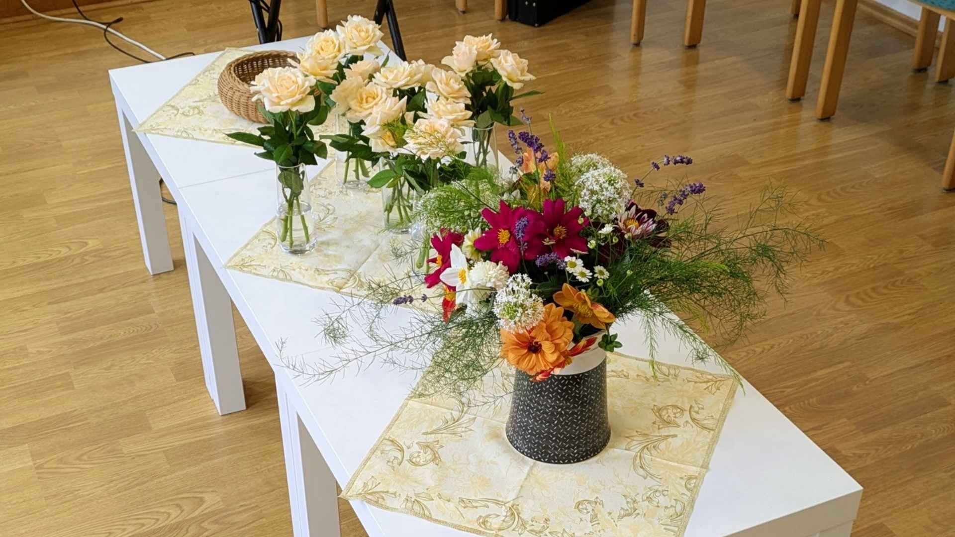 Mehrere Vasen stehen auf kleinen, weißen Beistelltischen in der Mitte eines Raumes. In den hinteren Vasen stehen zart cremefarbene Rosen, in der vorderen Vase steht ein Arrangement aus verschiedenen Dahlien und Gräsern.