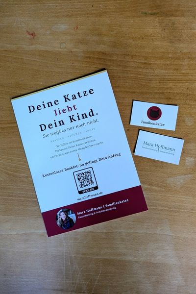 Auf einem Holztisch liegen ein A4-Plakat mit Werbung für "Familienkatze" von Mara Hoffmann, sowie passende Visitenkarte.