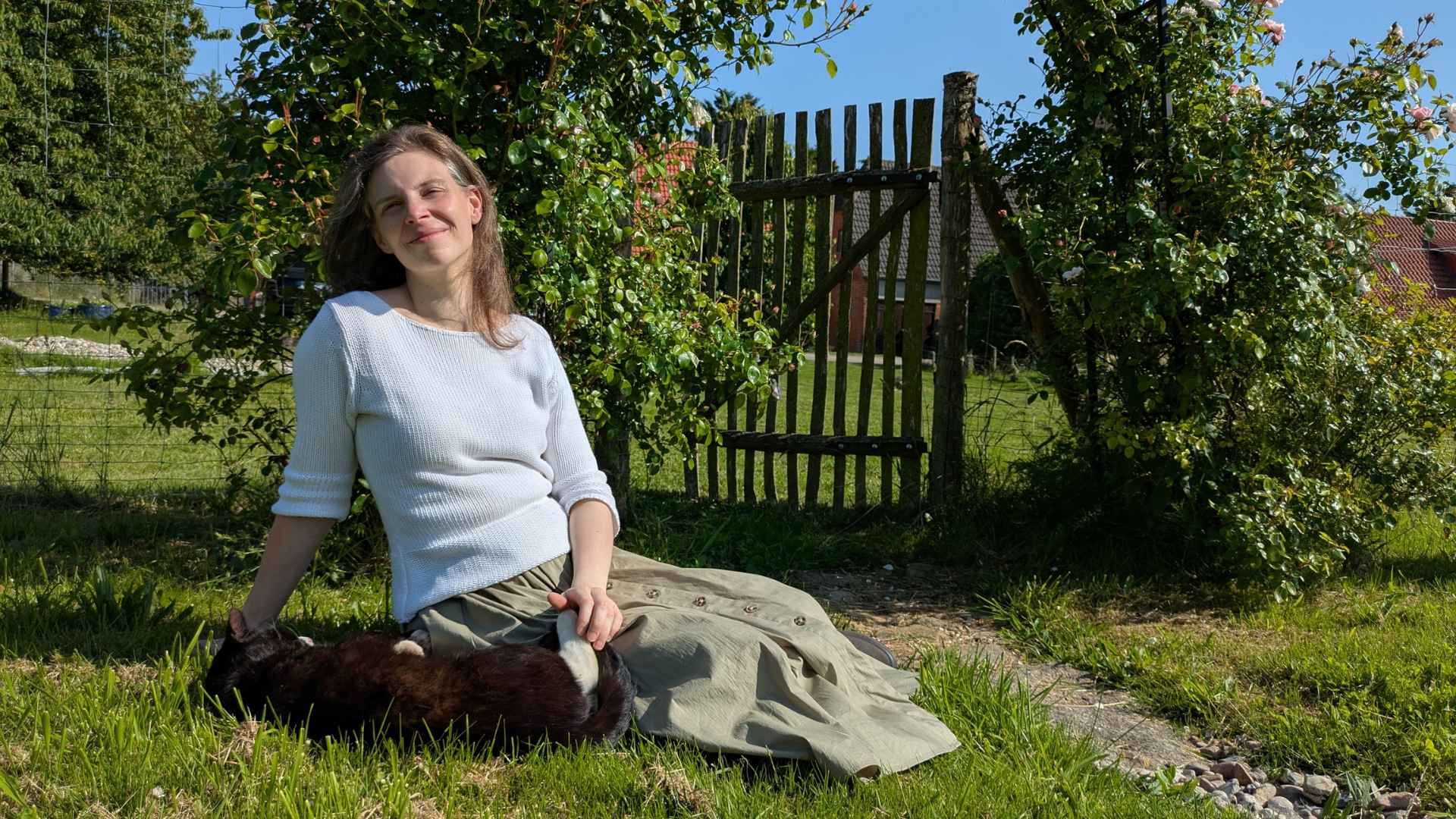 Eine Frau mit weißem Strickpullover und khaki-farbenen Rock sitzt auf einer Wiese vor einem Rosenbogen und krault einen schwarz-weißen Kater.