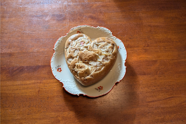 Auf einem Holztisch liegt auf einem alten Kuchenteller ein Apfelkuchen in Herzform.