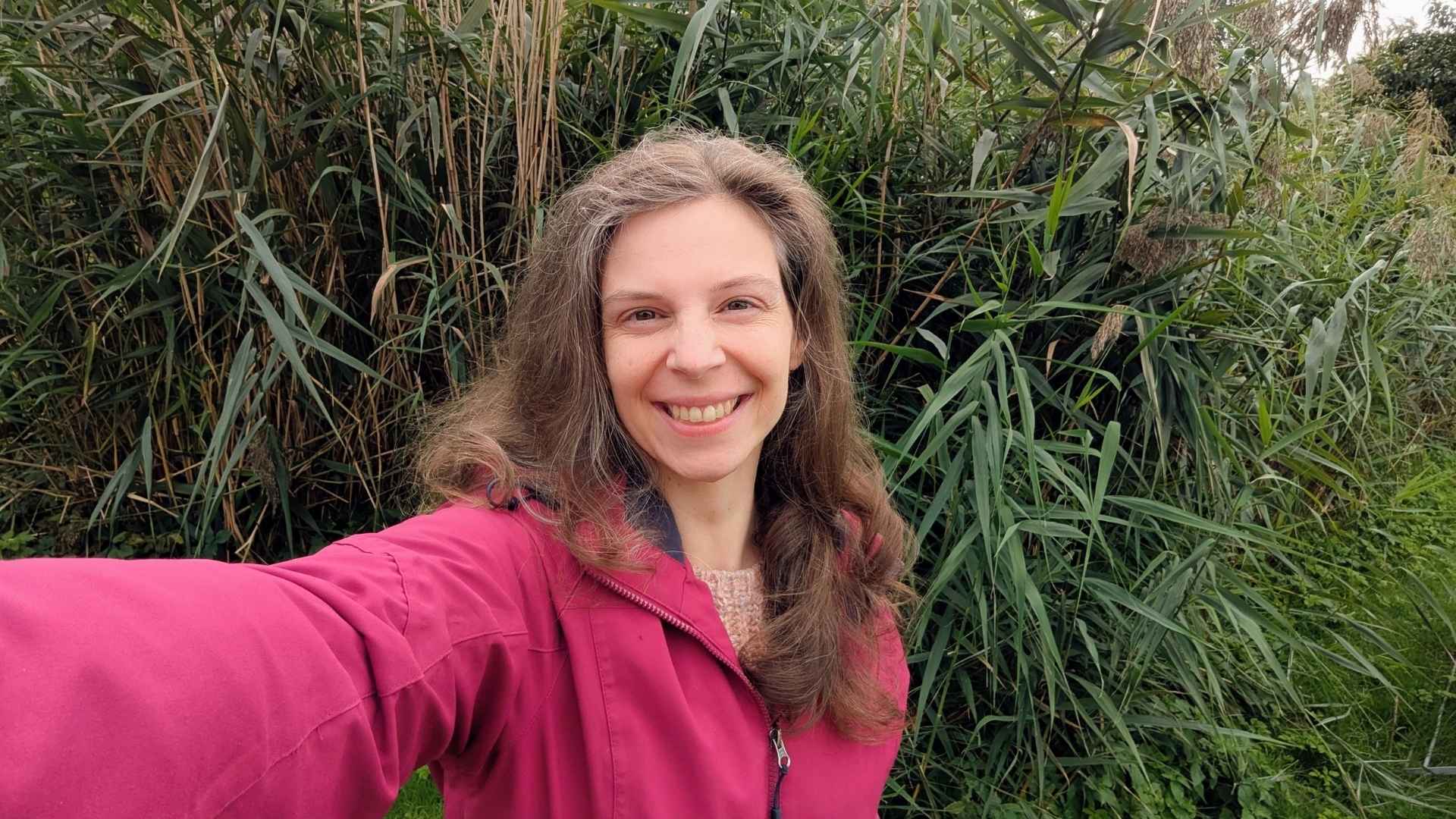 Eine junge Frau mit brünettem Haar und beerenfarbener Regenjacke steht vor einem Schilfbeet und knipst lachend ein Selfie.
