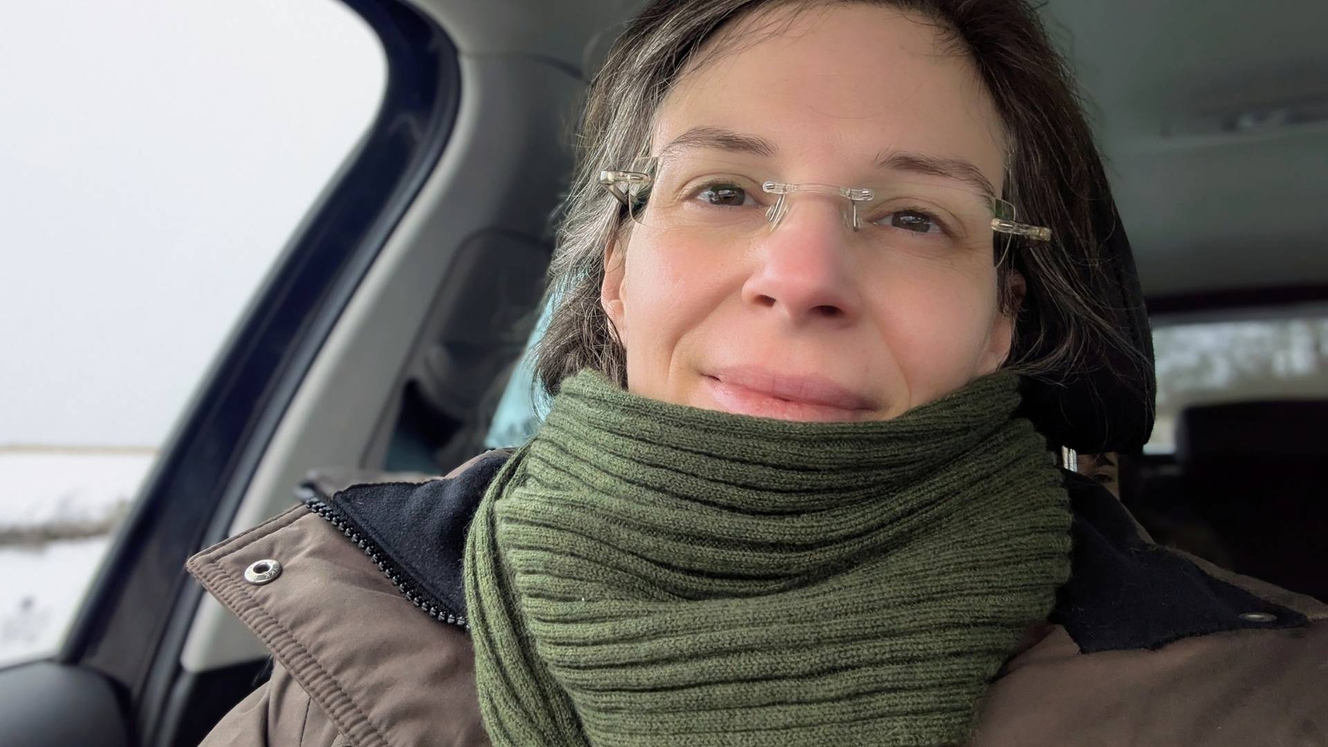 Eine junge Frau sitzt auf dem Beifahrersitz eines Autos. Sie ist winterfest mit einem braunen Parka und einem dunkelgrünem Schal eingemummt. Sie trägt eine randlose , klare Brille. Ihr Ausdruck ist müde, aber zuversichtlich. Sie lächelt ganz leicht.