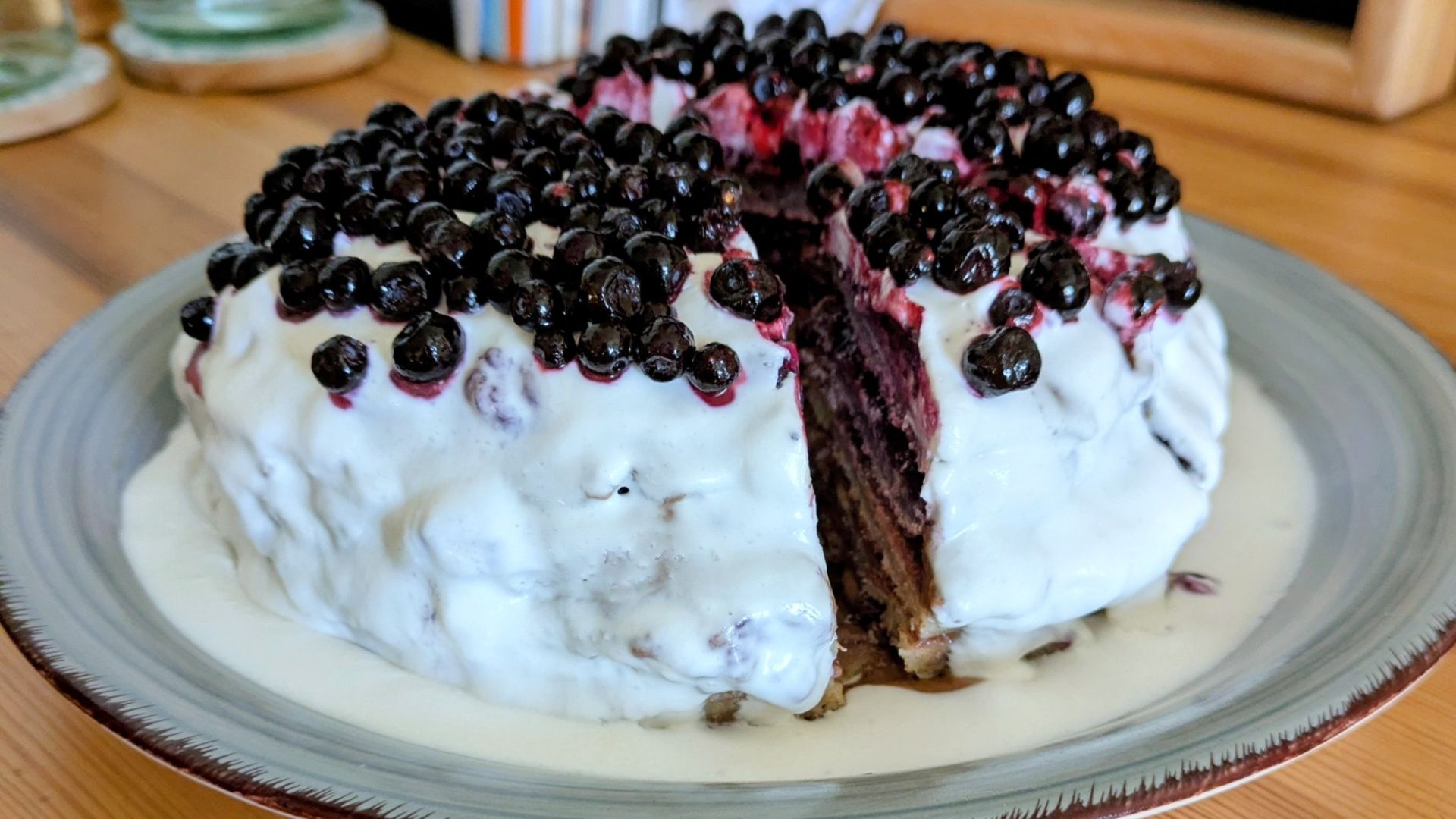 Eine Pfannkuchentorte nach Pettersson und Findus mit Carob-Creme und Heidelbeermarmelade in den Schichten und Kokossahne außen rum sowie Heidelbeeren obendrauf, ist auf einem grau-blauen Keramikteller angerichtet.