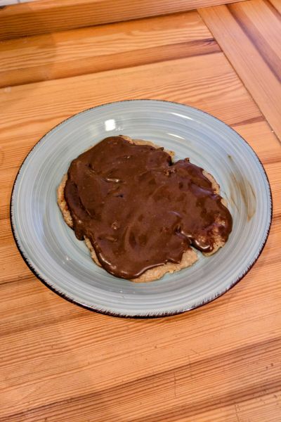 Auf einem grau-blauen Keramikteller liegt ein Pfannkuchen, der mit einer Carob-Creme bestrichen ist.
