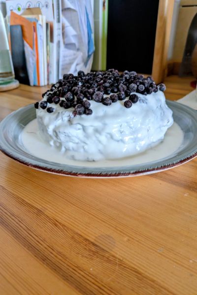 Die Pfannkuchentorte ist auf einem grau-blauen Keramikteller aufgeschichtet und nun mit weißer Kokossahne bestrichen und mit Heidelbeeren dekoriert.