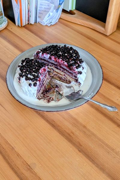 Dieselbe Pfannkuchentorte ist aufgeschnitten, ein Viertelsttück fehlt. Man kann direkt im Inneren die einzelnen Schichten der Torte sehen: sechs Pfannkuchen jeweils bestrichen mit einer Schicht Carob-Creme und einer Schicht Heidelbeermarmelade und ganz oben drauf sind frische Heidelbeeren gelegt.