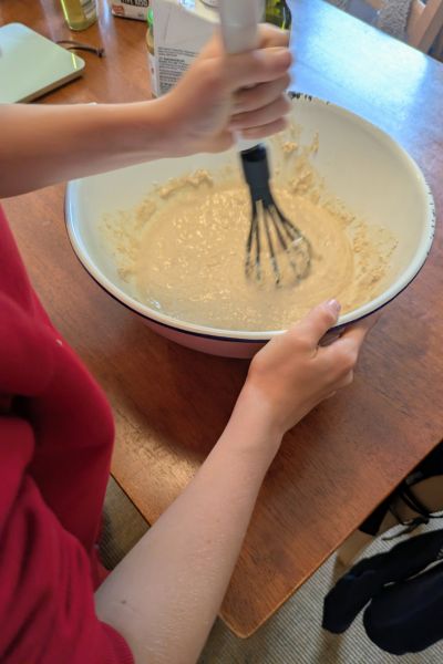 Ein Kind rührt mit einem Schneebesen Pfannkuchenteig in einer weißen Emailleschüssel an.