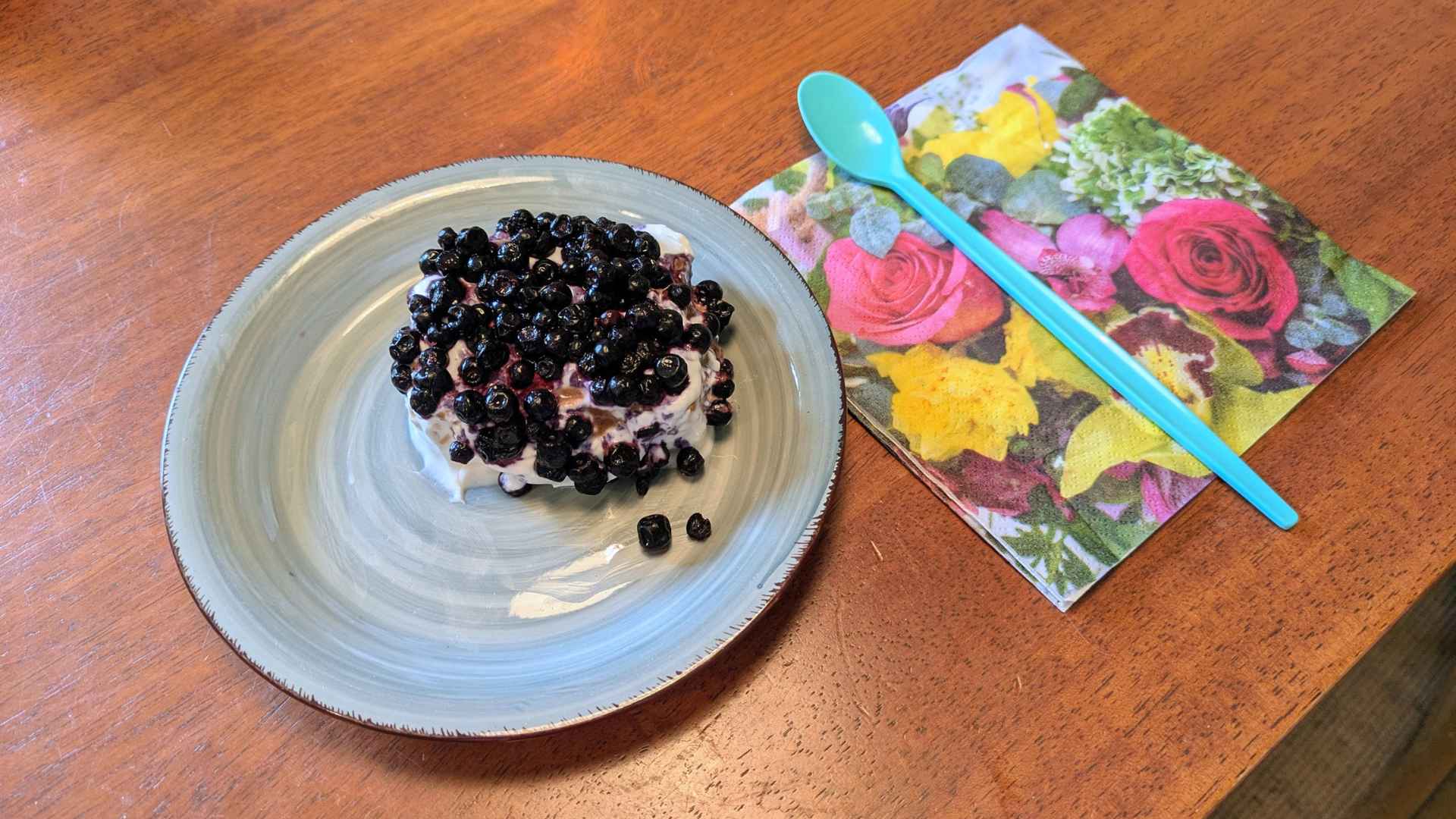 Auf einem blauen Keramikteller liegt ein Stück Kuchen: Pfannkuchen abwechselnd beschichtet mit Carobcreme und zuoberst Kokossahne und Heidelbeeren als Dekoration. Neben dem Teller liegt auf einer Serviette mit Rosenmotiv ein blauer Stiellöffel.