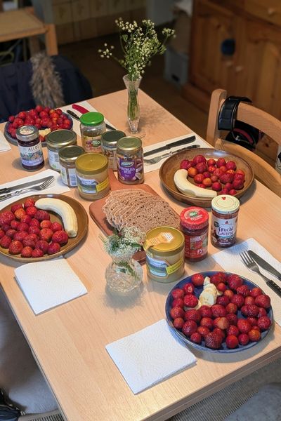 Auf einem Küchentisch stehen vier Teller voll mit Erdbeeren, dazu selbstgebackenes Brot auf einem Brettchen, Nussmuse und Marmelade. Ein Blumenstrauß aus Wiesenkräutern rundet den Sommer-Frühstückstisch ab.