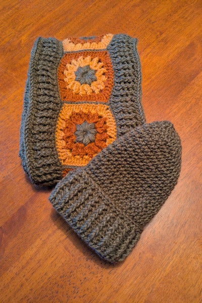 Auf einem Küchentisch liegt ein fertig gehäkeltes Set aus einer braunen Mütze und einem Schal mit rot-gelb.braunen Kokardenblumen, die von einer braunen Kante eingefasst sind.