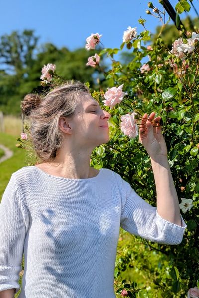 Eine Frau mit locker hochgestecktem Haar steht unter einem Rosenbogen und riecht an einer zart rosa farbenen Rose.