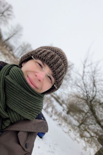 Eine junge Frau steht dick eingepackt in einem braunen Winterparka, brauner Mütze und grünem Schal vor einer Schneelandschaft. Die Kamera steht schräg, so dass das Bild insgesamt nach rechts gekippt ist.